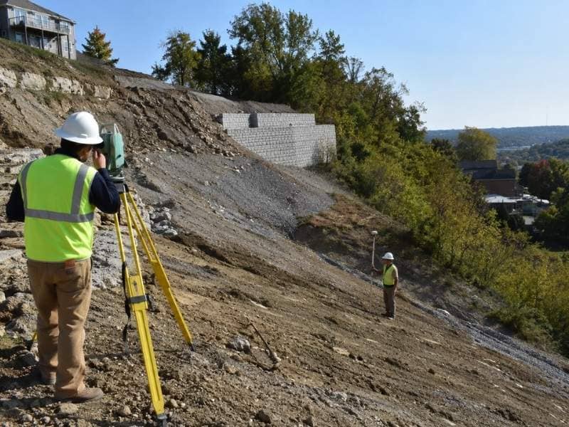 Survey crew collecting topographic elevation data across a steep Cincinnati construction site with retaining walls and exposed hillside soil